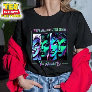 May include: Black t-shirt featuring a graphic of four villainous characters with vibrant green, purple, and blue tones. Text reads "WHO'S AFRAID OF LITTLE OLD ME - You Should Be". The shirt has a crew neck.