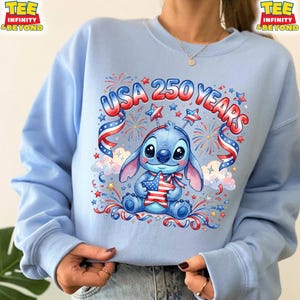 May include: Light blue sweatshirt featuring a cartoon character holding a star-shaped American flag. The design includes the text "USA 250 YEARS" with red, white, and blue accents, fireworks, and stars. The sweatshirt has long sleeves.