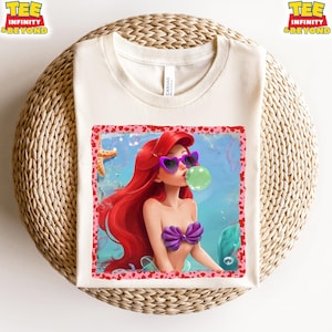 May include: Cream-colored t-shirt featuring a colorful illustration of a mermaid with red hair, wearing heart-shaped sunglasses and a purple seashell top, blowing a bubble. The design is framed with a pink border. The words "TEE INFINITY & BEYOND" are printed on the top left and right.