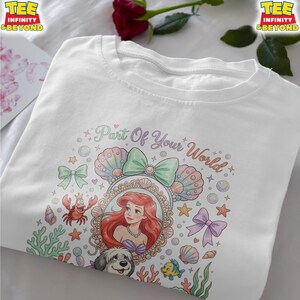May include: White t-shirt featuring a colorful graphic of Ariel from The Little Mermaid. The design includes the text "Part Of Your World", seashells, bows, and other aquatic elements. The shirt has a crew neck and short sleeves.