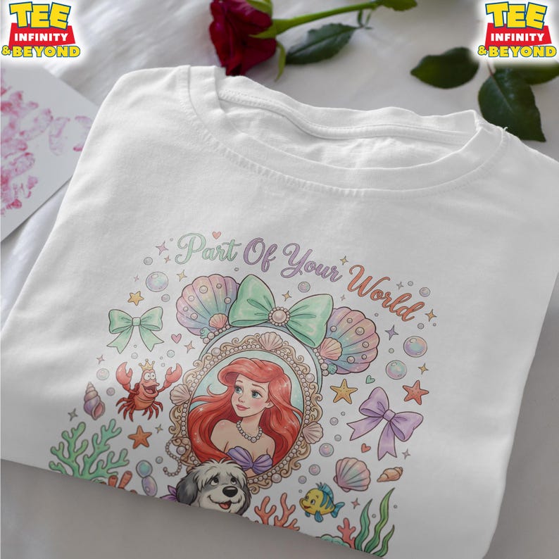May include: White t-shirt featuring a colorful graphic of Ariel from The Little Mermaid. The design includes the text "Part Of Your World", seashells, bows, and other aquatic elements. The shirt has a crew neck and short sleeves.