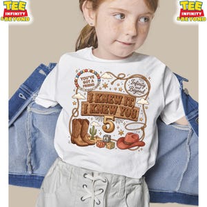 May include: A white t-shirt featuring a western-themed graphic with brown cowboy boots, a red cowboy hat, alphabet blocks, a cactus, and a lasso. The text reads: You've Got A Friend, Infinity and Beyond, I Knew It, I Knew You, and the number 5.