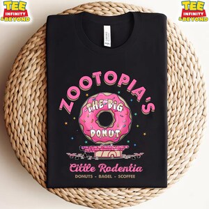 Puede incluir: Camiseta negra con un gr&aacute;fico de rosquilla rosa y el texto "Zootopia's The Big Donut" y "Little Rodentia". La camiseta tambi&eacute;n incluye las palabras "DONUTS - BAGEL - SCOFFEE". La camiseta se muestra sobre una superficie tejida.