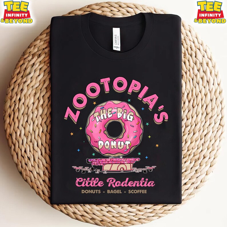 Puede incluir: Camiseta negra con un gr&aacute;fico de rosquilla rosa y el texto "Zootopia's The Big Donut" y "Little Rodentia". La camiseta tambi&eacute;n incluye las palabras "DONUTS - BAGEL - SCOFFEE". La camiseta se muestra sobre una superficie tejida.