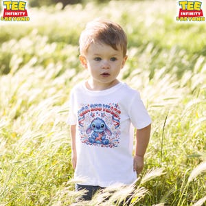 May include: A young child wearing a white t-shirt with a graphic of the character Stitch holding an American flag. The shirt also features the text "USA 250 YEARS" in red and blue. The child is standing in a field of tall grass.