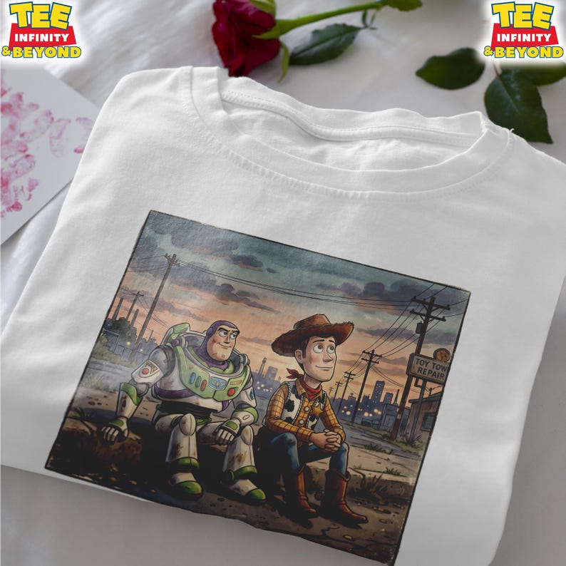 May include: White t-shirt with a watercolour illustration of Buzz Lightyear and Woody from Toy Story. The image shows the characters sitting together with a cityscape background. The shirt has the text "TEE INFINITY & BEYOND" at the top.