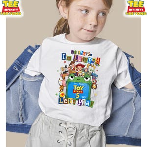 May include: A white short-sleeve t-shirt featuring a colorful Toy Story 5 graphic with a green frog character holding a sign that reads Toy Story 5. The text above says Hi there, I'm Lilypad and the text below says Let's Play, surrounded by characters.