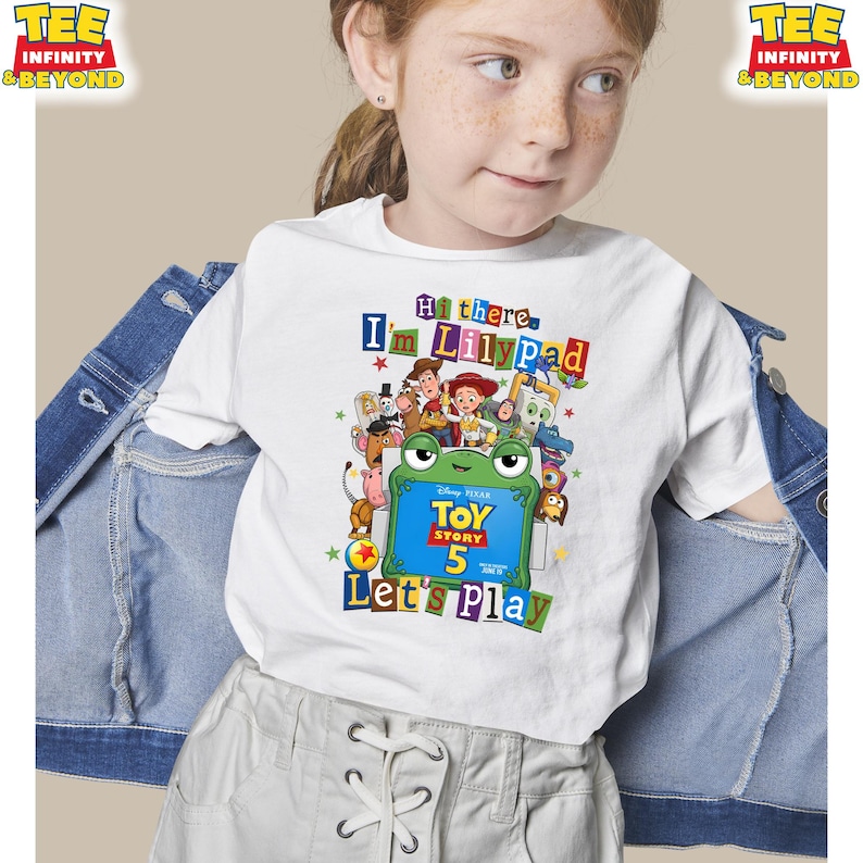 May include: A white short-sleeve t-shirt featuring a colorful Toy Story 5 graphic with a green frog character holding a sign that reads Toy Story 5. The text above says Hi there, I'm Lilypad and the text below says Let's Play, surrounded by characters.