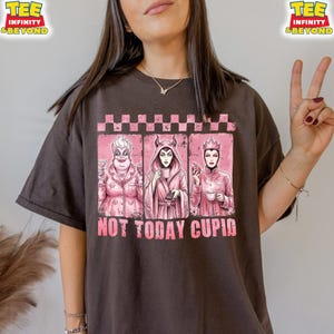 May include: Dark brown t-shirt featuring a pink graphic with three cartoon villains. The graphic includes the text "NOT TODAY CUPID" in pink. The shirt has a crew neck and short sleeves.