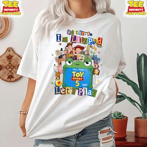 May include: A white t-shirt featuring a colorful graphic of Toy Story characters surrounding a green frog illustration. The text reads Hi there, I'm Lilypad, Toy Story 5, and Let's Play. The design includes various animated figures in a collage style.