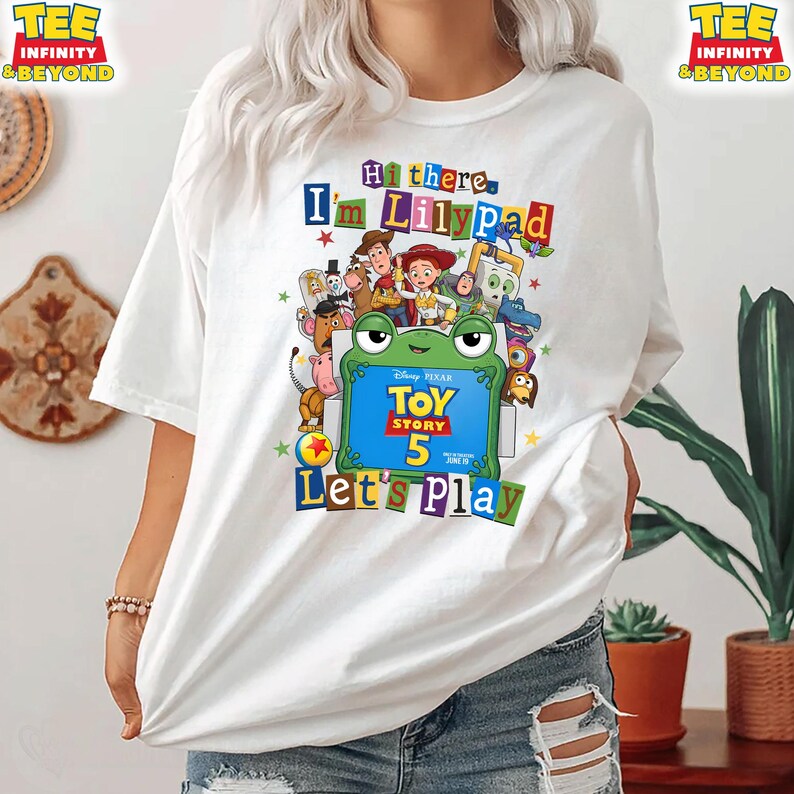 May include: A white t-shirt featuring a colorful graphic of Toy Story characters surrounding a green frog illustration. The text reads Hi there, I'm Lilypad, Toy Story 5, and Let's Play. The design includes various animated figures in a collage style.