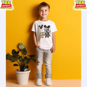 May include: A white short-sleeved t-shirt featuring a graphic print of a cartoon Mandalorian character and a small green creature, both wearing mouse-ear headbands. The Mandalorian holds a black balloon shaped like mouse ears.