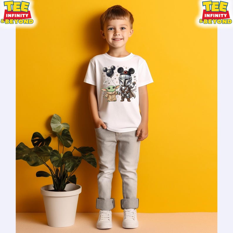May include: A white short-sleeved t-shirt featuring a graphic print of a cartoon Mandalorian character and a small green creature, both wearing mouse-ear headbands. The Mandalorian holds a black balloon shaped like mouse ears.