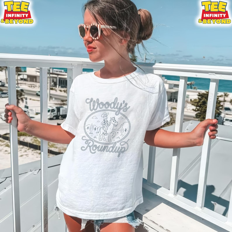 May include: A white short-sleeve t-shirt featuring a dark gray graphic print of a cowboy on a horse inside a rope-style oval frame. The text "Woody's Roundup" is written in a cursive font above and below the illustration of the cowboy.
