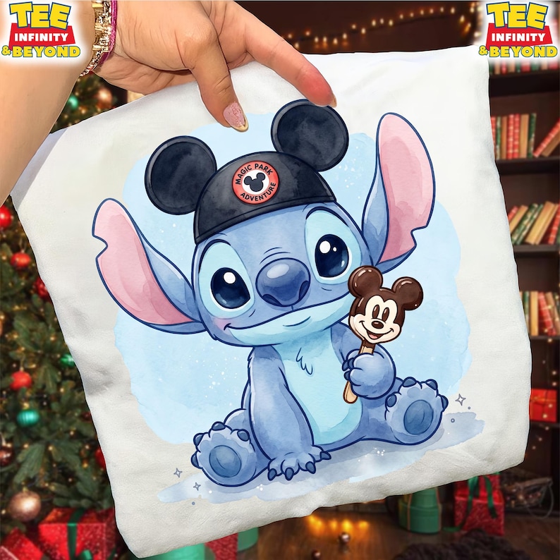 K&ouml;nnte beinhalten: Wei&szlig;es Kissen mit einer blauen Cartoonfigur, die einen schwarzen Hut mit dem Text "Magic Park Adventure" tr&auml;gt. Die Figur h&auml;lt einen Mickey Mouse Eis am Stiel. Das Kissen hat einen hellblauen Aquarellhintergrund.