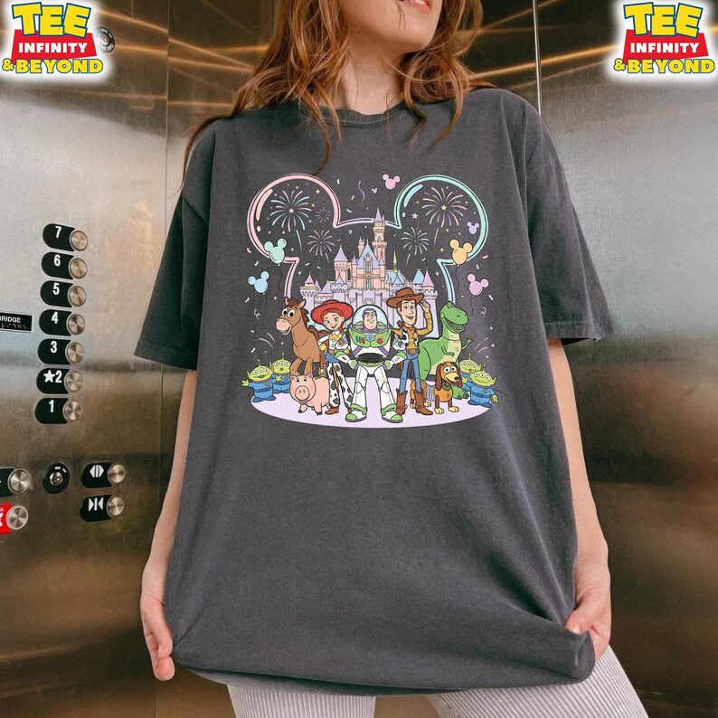 May include: Dark gray t-shirt featuring a colorful graphic of Disney's Toy Story characters in front of a castle with fireworks. The design includes Buzz Lightyear, Woody, and other characters. The shirt has the text "TEE INFINITY & BEYOND" on the top corners.
