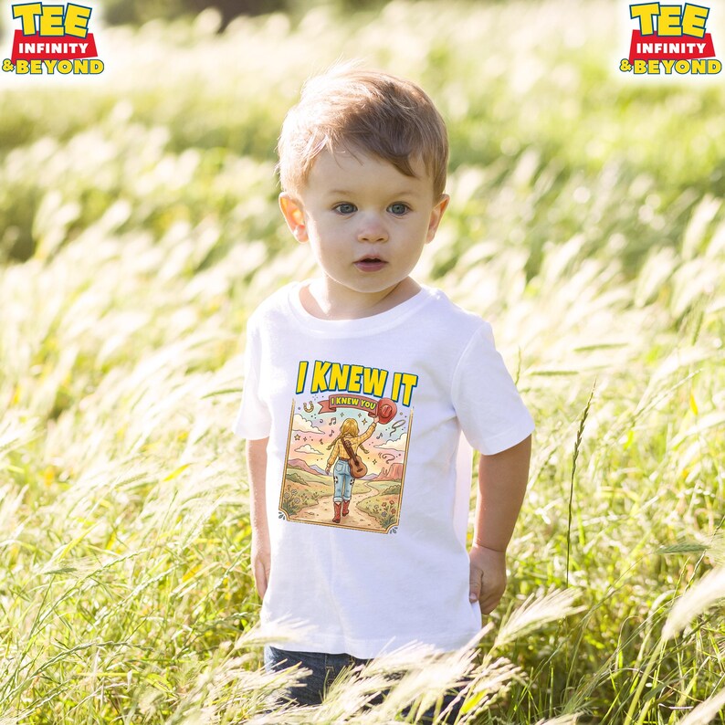 Puede incluir: Una camiseta blanca de manga corta con un estampado gr&aacute;fico colorido de una persona caminando por un sendero en un paisaje des&eacute;rtico. El texto "I KNEW IT" est&aacute; impreso en letras amarillas grandes sobre la ilustraci&oacute;n.