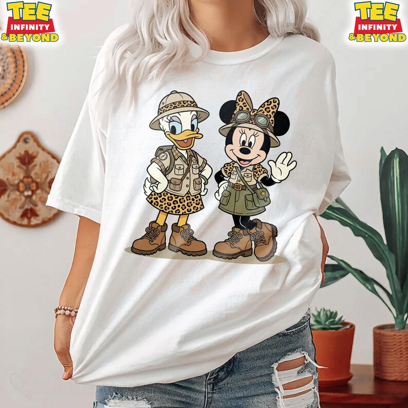 May include: White t-shirt featuring cartoon characters Daisy Duck and Minnie Mouse dressed in safari outfits. Daisy Duck wears a khaki safari jacket and leopard print skirt. Minnie Mouse wears a similar outfit with a green skirt. The shirt has the text "TEE INFINITY & BEYOND".