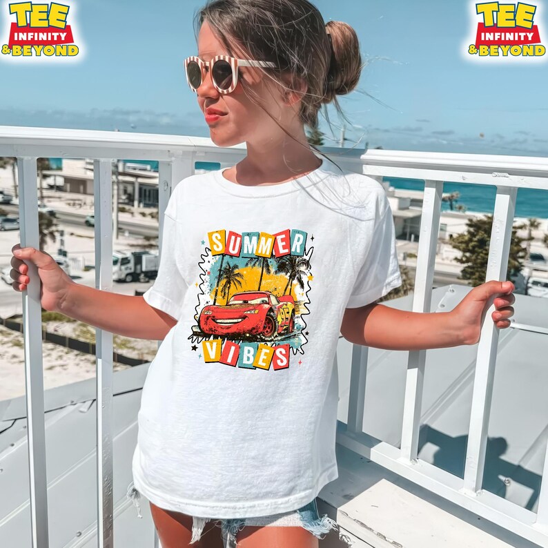 May include: White t-shirt featuring a graphic of a red race car with the text "SUMMER VIBES" in colorful letters. The design includes palm trees and a blue sky. The shirt is worn by a person wearing sunglasses.