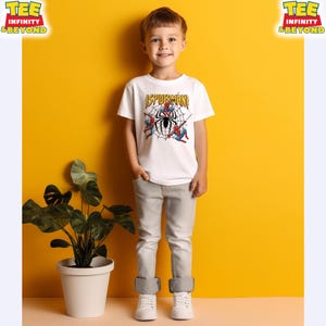 May include: A young child wearing a white t-shirt with a Spider-Man graphic. The shirt features the word "SPIDERMAN" in yellow, with a black spider and web design. The child is wearing gray pants and white sneakers, standing in front of a yellow background.