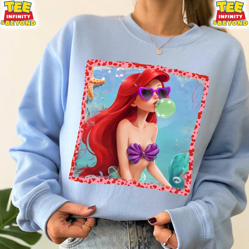 May include: Light blue sweatshirt featuring a graphic of a cartoon mermaid with red hair, heart-shaped sunglasses, and a purple seashell top. The mermaid is blowing a bubble. The sweatshirt has the text "TEE INFINITY & BEYOND" on the top left and right.