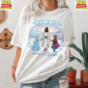 以下が含まれることがあります： 「LET IT GO AND LET JESUS」のテキストが入った、カラフルなグラフィックが特徴の白いTシャツ。グラフィックは、イエスが2人の女の子と手をつないでいる冬のシーンを描いています。
