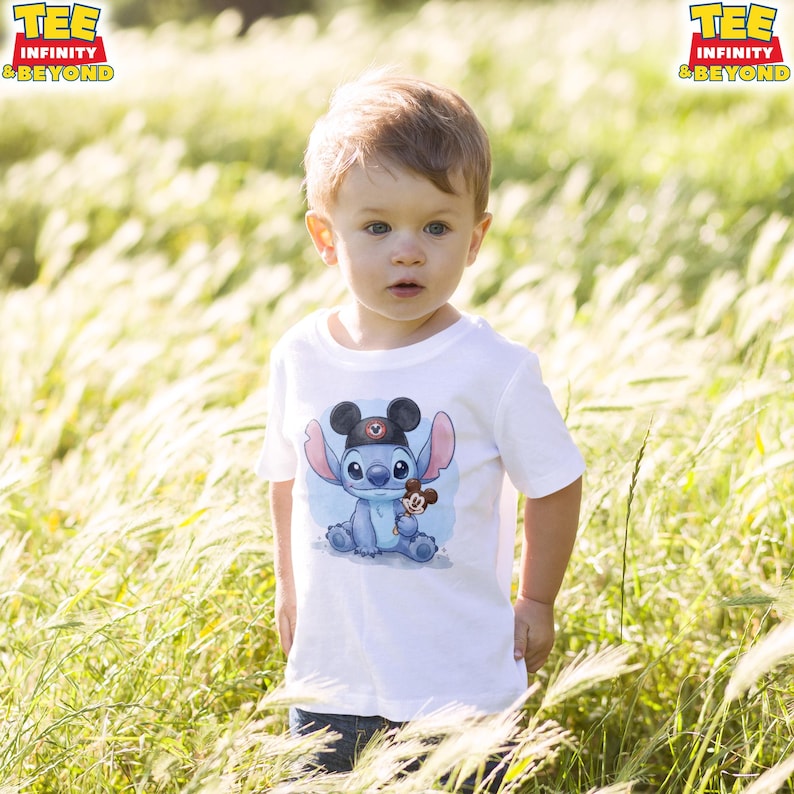 K&ouml;nnte beinhalten: Ein Kind tr&auml;gt ein wei&szlig;es T-Shirt mit einer Cartoon-Illustration von Stitch und Mickey Mouse. Auf dem T-Shirt steht der Text "TEE INFINITY & BEYOND" in einem gelb-roten Logo. Das Kind steht in einem Feld mit hohem Gras.