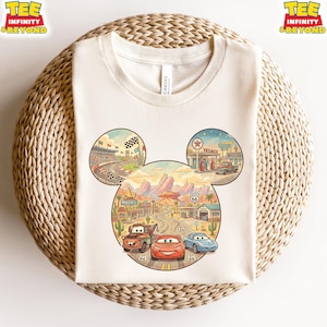 May include: Cream-colored t-shirt featuring a colorful graphic of the Cars movie characters in a Mickey Mouse head shape. The design includes race tracks, desert landscapes, and Route 66 elements. The shirt has the text "TEE INFINITY & BEYOND".
