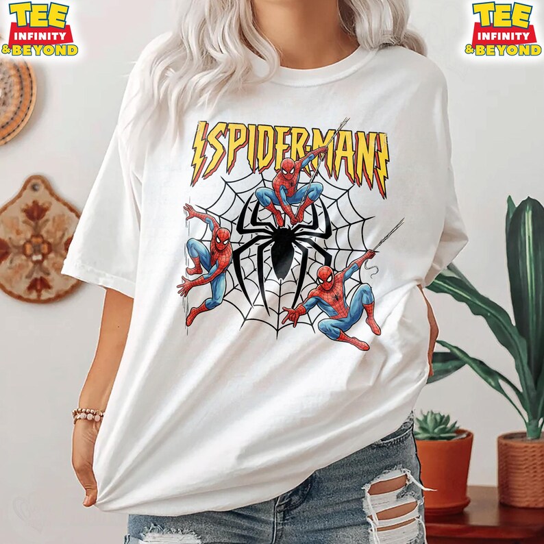 May include: White t-shirt featuring a graphic of Spider-Man in a web with the text "SPIDERMAN!" in yellow and red. The design includes three Spider-Man figures in action poses, with a large black spider in the center.