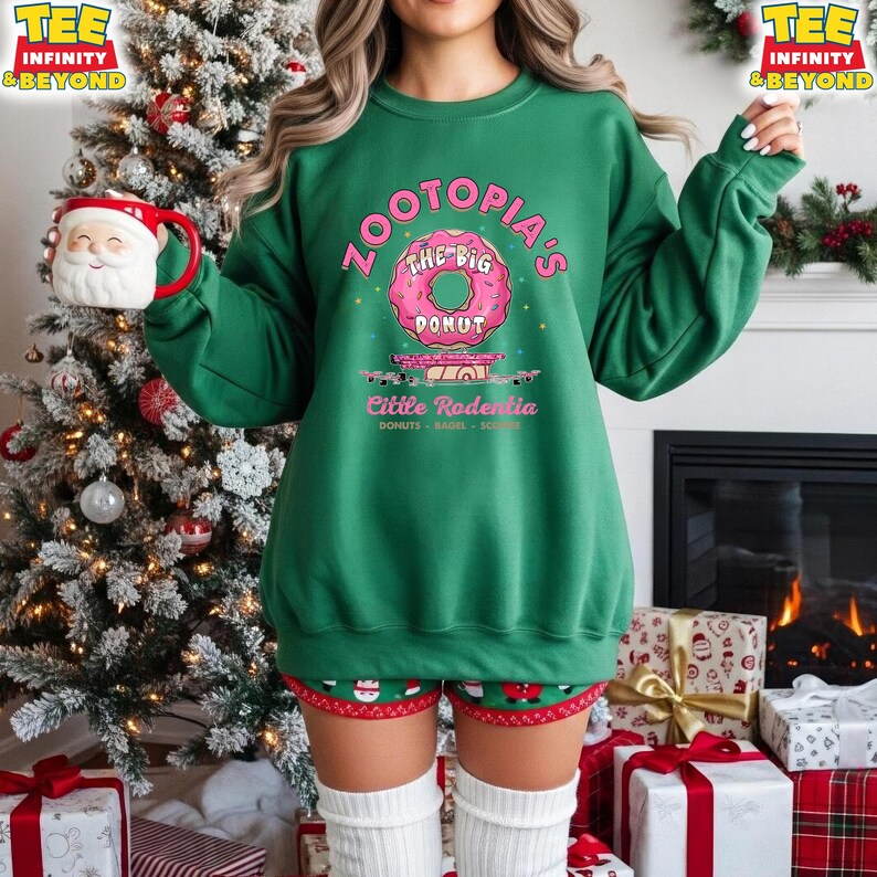 Puede incluir: Una sudadera verde con el texto "Zootopia's The Big Donut" y "Little Rodentia" en rosa. La sudadera se combina con pantalones cortos con tem&aacute;tica navide&ntilde;a y calcetines blancos. Una taza de Pap&aacute; Noel se sostiene cerca de un &aacute;rbol de Navidad decorado.