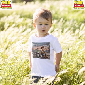 May include: A white t-shirt with a graphic of Buzz Lightyear and Woody from Toy Story. The shirt is worn by a young child in a field of tall grass. The shirt has the text "TEE INFINITY & BEYOND" in the upper left and right corners.