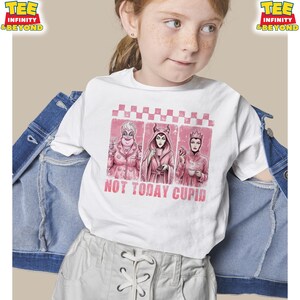 May include: White t-shirt featuring a pink graphic with three cartoon villains and the text "NOT TODAY CUPID". The shirt has a crew neck and short sleeves. The shirt is being worn by a person.