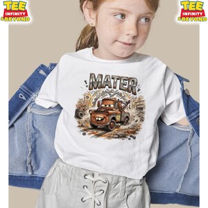 May include: White t-shirt featuring a cartoon image of Mater from the Cars movie. The design includes the name "MATER" above the character, with a brown tow truck surrounded by gears and debris. The shirt is worn by a young person.
