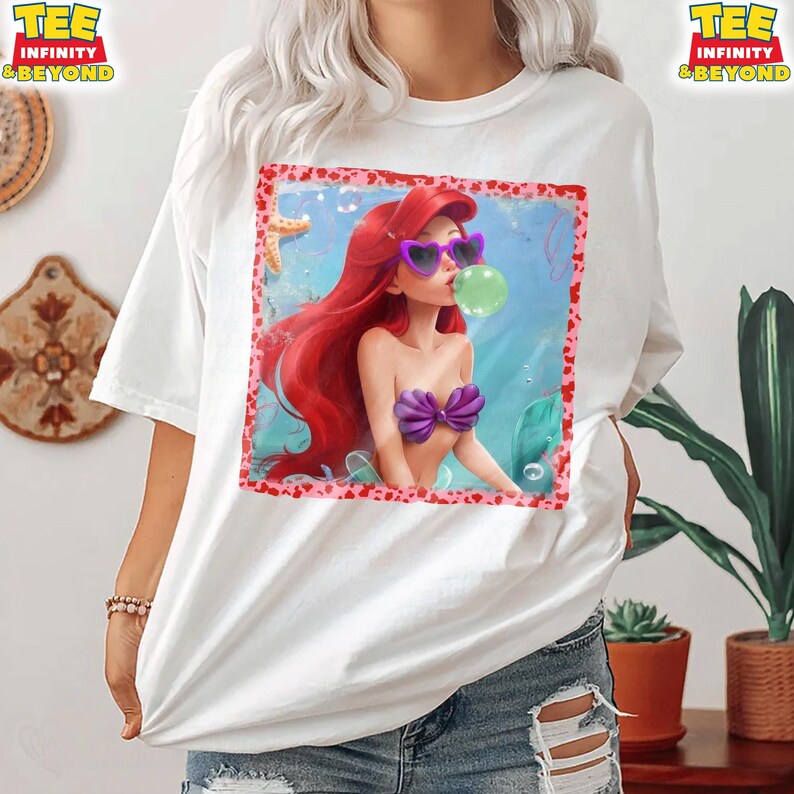 May include: White t-shirt featuring a graphic of a cartoon mermaid with red hair, wearing heart-shaped sunglasses and a purple seashell top, blowing a bubble. The graphic is framed with a pink border. The shirt has the text "TEE INFINITY & BEYOND" on the top.