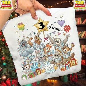 Puede incluir: Camiseta blanca con una ilustraci&oacute;n de dibujos animados de animales celebrando un cumplea&ntilde;os. El dise&ntilde;o incluye un globo de estrella dorada con el n&uacute;mero 3, una tarta y la palabra "Name". La camiseta est&aacute; decorada con globos y cajas de regalo.