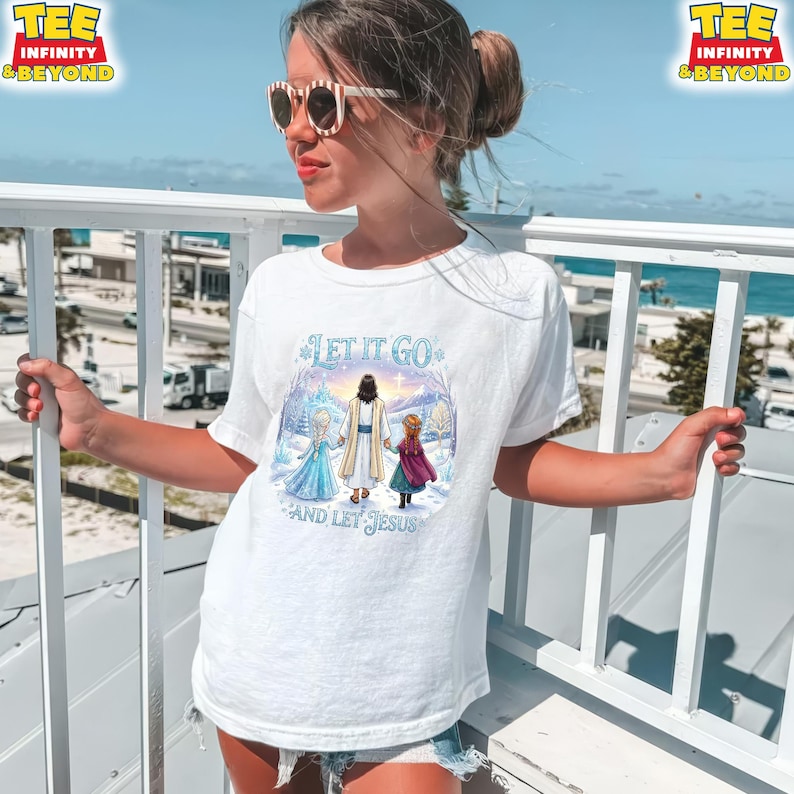 以下が含まれることがあります： 「LET IT GO AND LET JESUS」の文字が入った白いTシャツ。冬のシーンでイエスが子供たちと手をつないでいるグラフィック。Tシャツは半袖で、クルーネックです。