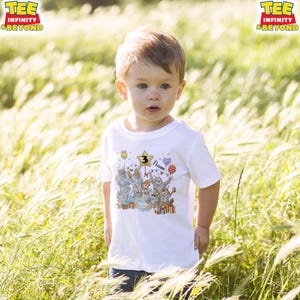 Puede incluir: Una camiseta blanca con un gr&aacute;fico colorido de animales de dibujos animados, globos y regalos. El n&uacute;mero "3" se muestra en una estrella dorada, con la palabra "Name" debajo. La camiseta la lleva un ni&ntilde;o peque&ntilde;o en un campo de hierba alta.
