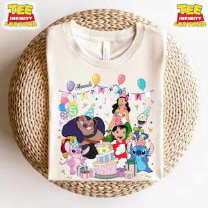 May include: A cream-coloured t-shirt with a vibrant graphic of animated characters, a birthday cake, and balloons. The text "Amanda Birthday" is above the characters. The shirt has the text "TEE INFINITY & BEYOND" on the top left and right.