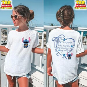 May include: White t-shirt featuring a blue cartoon character with large ears and a colorful flower design on the front. The back of the shirt has a blue heart with the text "everyday is autism awareness in my world."
