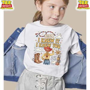 May include: A white t-shirt featuring a graphic print of a cowgirl with a red hat, cowboy boots, a sheriff badge, and alphabet blocks. The text reads "YOU'VE GOT A FRIEND IN ME", "I KNEW IT I KNEW YOU", and "It's a Jessie Story Baby Just Say Yes".