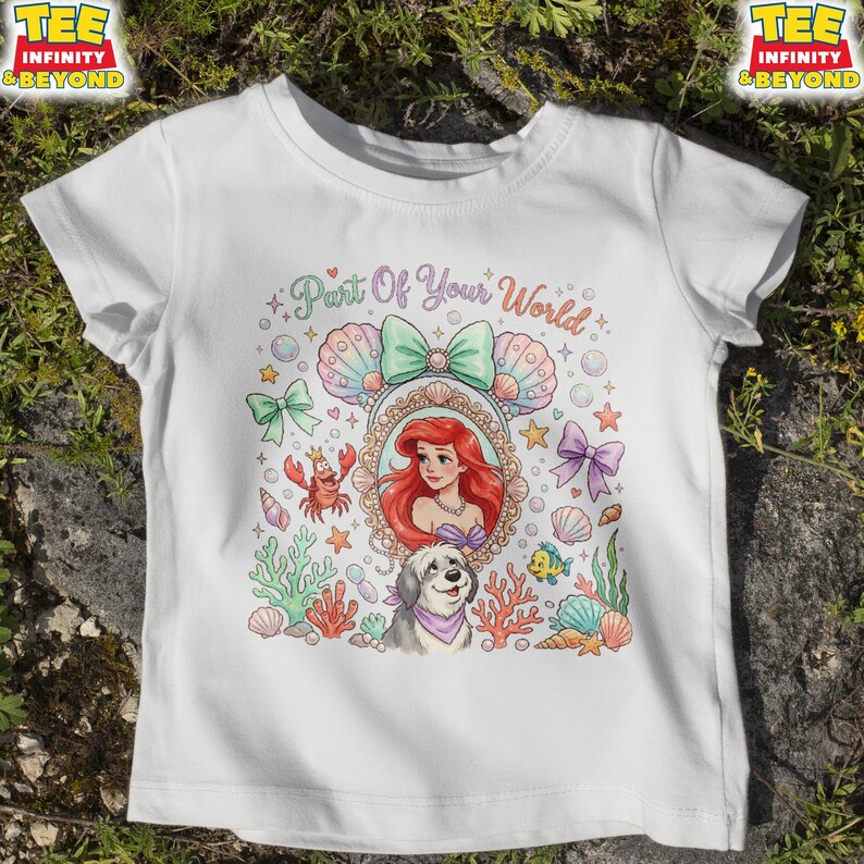 May include: White t-shirt featuring a colorful illustration with the text "Part Of Your World." The design includes a portrait of a red-haired woman, a dog, seashells, and other sea-themed elements. The shirt has short sleeves.