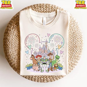 May include: Cream-colored t-shirt featuring a colorful illustration of Disney's Toy Story characters in front of a castle. The design includes fireworks, balloons, and the text "TEE INFINITY & BEYOND" in yellow and orange.