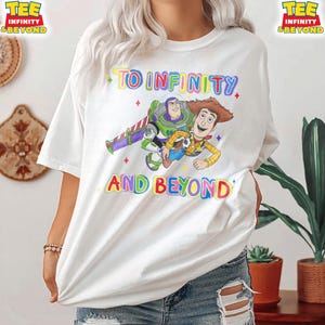 May include: White t-shirt with a colourful graphic of Buzz Lightyear and Woody from Toy Story, and the words "TO INFINITY AND BEYOND". The tee has a crew neck and short sleeves.