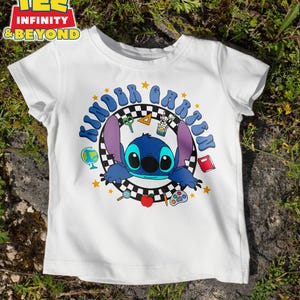 May include: White t-shirt featuring a blue cartoon character with large ears and the text "Kindergarten" in blue. The design includes stars, school supplies, and a checkered background. The shirt has short sleeves and a crew neck.