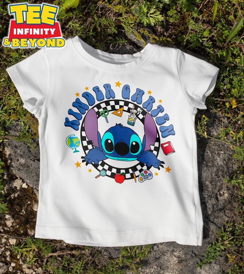 May include: White t-shirt featuring a blue cartoon character with large ears and the text "Kindergarten" in blue. The design includes stars, school supplies, and a checkered background. The shirt has short sleeves and a crew neck.