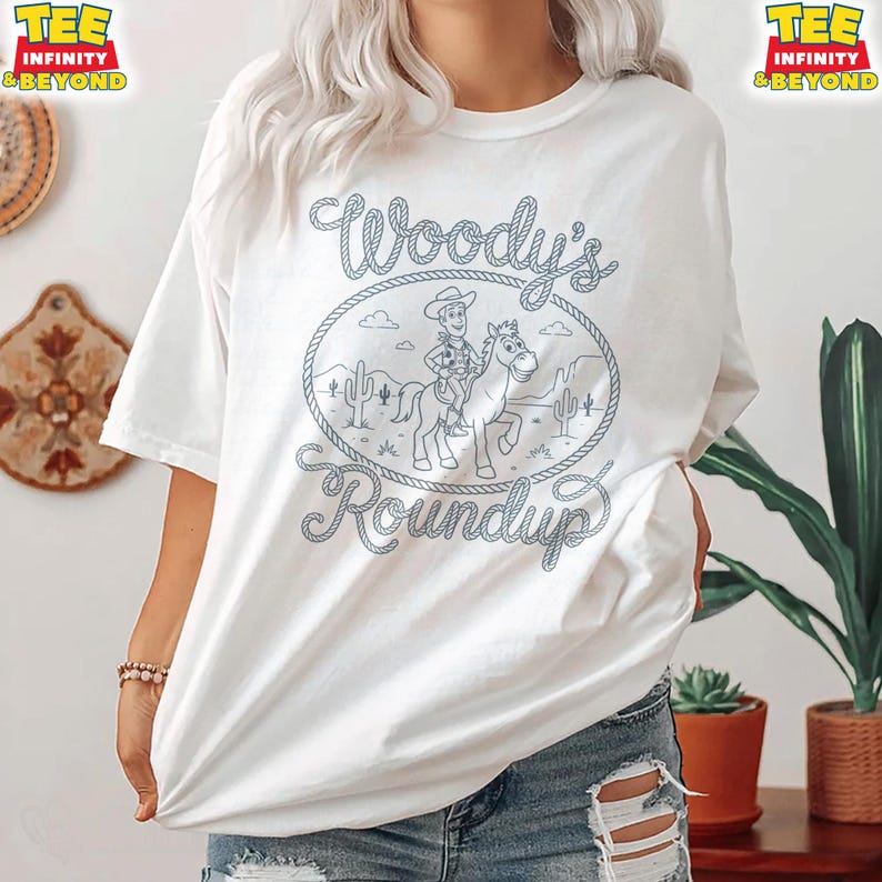 May include: A white short-sleeve t-shirt featuring a blue line-art graphic of a cowboy riding a horse inside a rope-style oval frame. The text Woody's Roundup is printed in a cursive font around the frame, with desert cacti in the background.