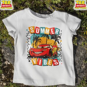 May include: White t-shirt featuring a graphic of a red race car with the number 95, palm trees, and the words "SUMMER VIBES" in colorful block letters. The shirt has short sleeves and a crew neck.
