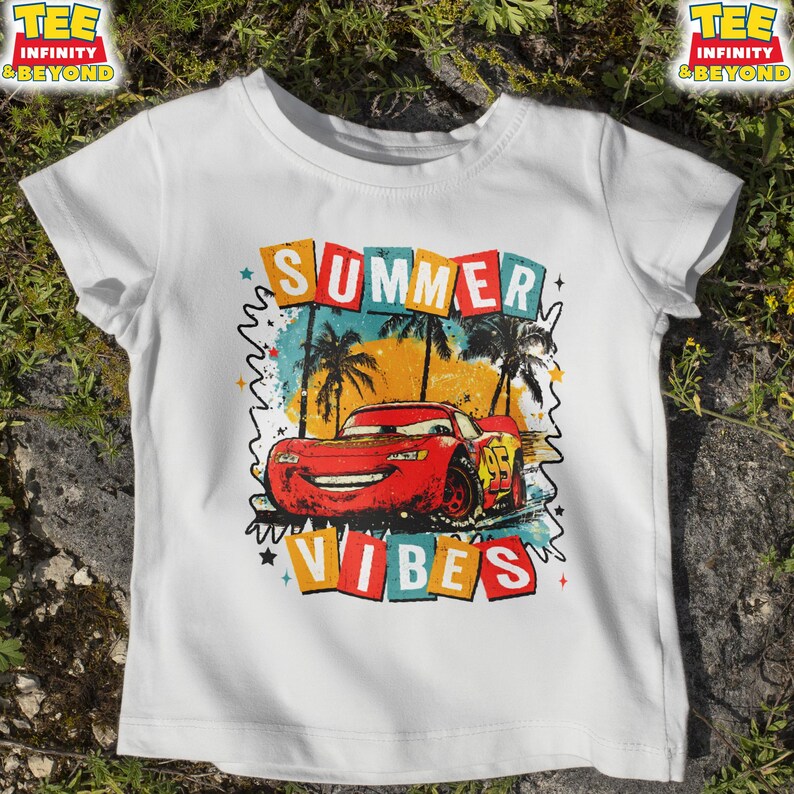 May include: White t-shirt featuring a graphic of a red race car with the number 95, palm trees, and the words "SUMMER VIBES" in colorful block letters. The shirt has short sleeves and a crew neck.