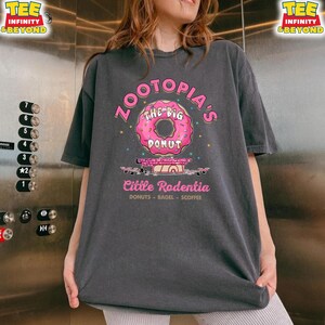 Puede incluir: Camiseta gris oscuro con un gr&aacute;fico de rosquilla rosa y el texto "Zootopia's The Big Donut" y "Cittle Rodentia". La camiseta tambi&eacute;n tiene las palabras "Donuts - Bagel - Scoffee" impresas debajo.
