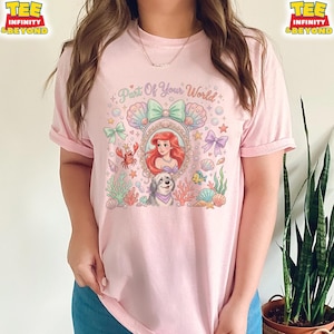 May include: A light pink t-shirt featuring a colorful graphic of Ariel from The Little Mermaid. The design includes the text "Part Of Your World", a portrait of Ariel, a dog, and various sea-themed elements like seashells and coral.